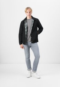 Veste noire avec intérieur à motifs, t-shirt gris, jeans à lavage clair et baskets blanches. Présente un devant boutonné et une coupe décontractée.