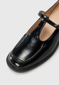 Glossy black leather Mary Jane shoe with a T-strap and beige interior lining, shown in close-up on a white background.