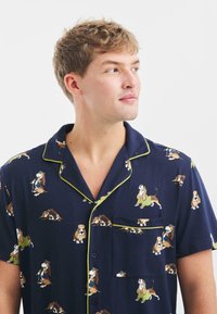 La camisa de pijama de manga corta para hombres en color azul marino presenta un estampado de perro basset, acentos en verde, un bolsillo en el pecho y un diseño clásico de botones.