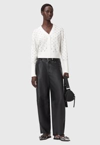 Weißer gestrickter Cardigan mit Sternmustern, schwarze Leder-Hosen im weit geschnittenen Stil und gemusterte Sandalen. Tote-Bag in der Hand, neutraler Hintergrund.