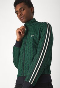 Zeleni zip-up jakna s geometrijskim uzorkom, bijelim prugama na rukavima, rebrastim manžetama i malim logom na prsima.