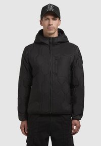 Schwarze Daunenjacke mit hexagonalen Polstern, Reißverschluss und abnehmbarem Kapuzen. Verfügt über eine Brusttasche und strapazierfähigen Stoff. Begleitet von einer schwarzen Mütze.