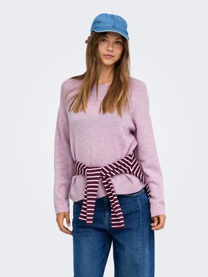Jeune femme portant une casquette bleue, un pull violet clair, un jean bleu et une chemise rayée bordeaux nouée autour de sa taille sur un fond uni.