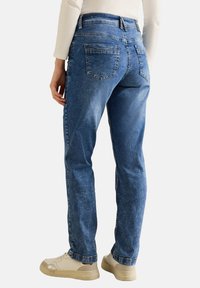 Jeans de mezclilla azul claro con corte recto, con bolsillos traseros y un desgaste mínimo. Combinados con una blusa blanca de manga larga y zapatillas beige.