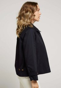 Veste bleu marine en tissu léger, avec un col, des poches avant et des accents de boutons marron. Coupe droite avec des manches longues.