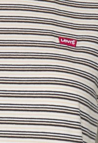 Pruhované tričko v krémové, černé a oranžové barvě; na hrudi má červenou nášivku s logem Levi's. Materiál vypadá měkký a texturovaný.