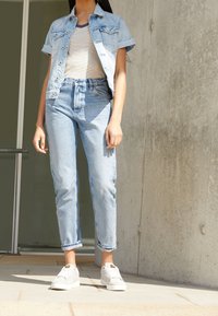 Femme portant une veste en denim, une chemise rayée, un jean bleu clair et des baskets blanches, se tenant à l'extérieur à côté d'un mur en béton.