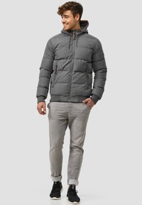 Grå puffjakke med hette og glidelås. Har to glidelomme og teksturert stoff. Kombinert med lys grå bukser og svarte joggesko.