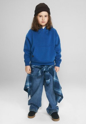 Dziecko ubrane w czarną czapkę, niebieską bluzę z kapturem, zawiązaną koszulę w kratę wokół talii, luźne dżinsy i czarne trampki, stojące na jednolitym tle.