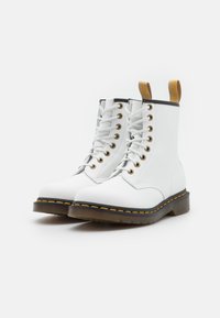 Botines de cuero blancos hasta el tobillo con puntera redondeada, ribetes negros, ojetes dorados y una lengüeta amarilla. Suela de goma con detalle de costura amarilla.