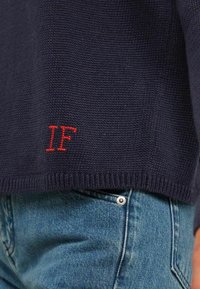 Pull en tricot bleu marine avec un ourlet côtelé, orné d'un détail brodé "IF" rouge en bas. Porté sur un jean bleu clair avec un bouton en métal argenté.