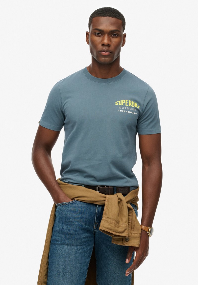 Camiseta de algodón azul para hombre con mangas cortas y cuello redondo. Presenta un logo amarillo "Superdry Outdoor" en el pecho. Se lleva con unos vaqueros y una chaqueta marrón atada a la cintura.
