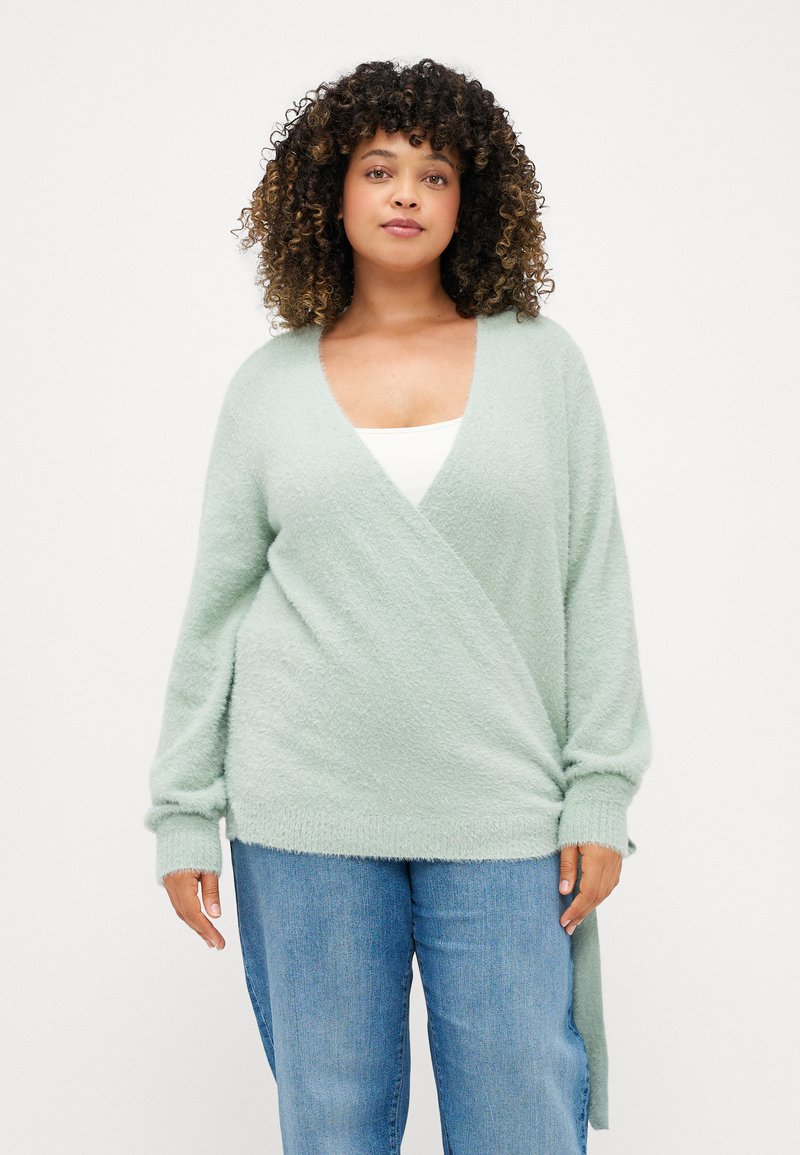 Ανοιχτό πράσινο, υφασμάτινο ζακέτα με μακριά μανίκια, V-κοπή, και λεπτομέρεια με λουράκι στο πλάι, συνδυασμένη με casual μπλε τζιν.