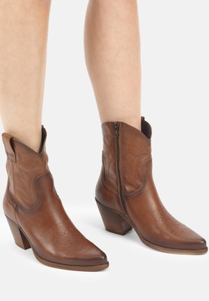 Braune Cowboy-Stiefel aus Leder, mittelhoch bis zur Wade, mit seitlichen Reißverschlüssen, getragen an nackten Beinen vor einem einfarbigen weißen Hintergrund.