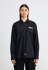 Camicia nera con bottoni, dotata di due tasche frontali e logo in beige. Realizzata in tessuto liscio e leggero; presenta maniche lunghe e colletto.