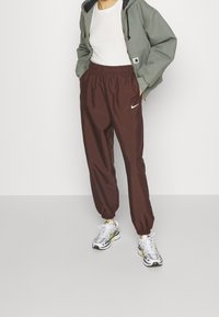 Hnědé jogger kalhoty s elastickým pasem a logem Nike, doplněné světle šedou mikinou, bílým tričkem a šedými teniskami se žlutými akcenty.