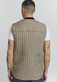 Beige quiltet vest med horisontale og vertikale sømmer, med krage og en glatt tekstur, båret over en hvit skjorte.