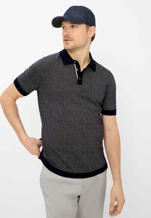 Mann trägt ein dunkelgraues Polohemd mit schwarzem Kragen und Bündchen, hellgraue Hosen und eine dunkelblaue Baseballkappe, schaut nach links.