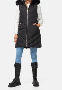 Veste sans manches noire matelassée en chevrons, capuche bordée de fourrure, fermetures éclair dorées et poches latérales ; portée avec un jean bleu et des bottes noires montantes jusqu'aux genoux.