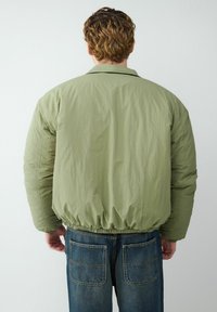 Jasnozielona kurtka puchowa z elastycznym dołem i luźnym krojem. Charakteryzuje się gładką, matową fakturą i klasycznym kołnierzem. Zestawiona z ciemnoniebieskimi dżinsami.