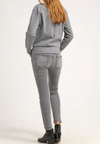 Donna con capelli lunghi, che indossa una felpa grigia, jeans skinny grigi e stivaletti neri alla caviglia, in piedi con le mani sui fianchi, rivolta di spalle verso l'osservatore.