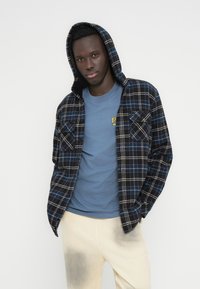 Πουκάμισο φανέλας με καρό μοτίβο σε σκούρο μπλε και ανοιχτό γκρι, με κουκούλα και τσέπες στο στήθος. Συνδυασμένο με μπλε μπλουζάκι και ανοιχτόχρωμο παντελόνι.