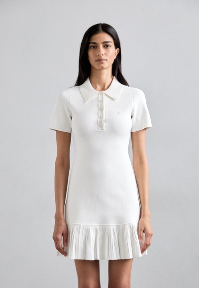 Robe tricotée blanche avec col polo, manches courtes, patte de boutonnage et ourlet plissé. Matériau texturé et détail de logo subtilement brodé.
