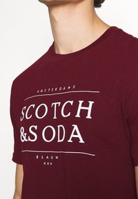 Bourgogne bomulds t-shirt med hvid tekst, der står "SCOTCH & SODA AMSTERDAMS BLAUW," med rund hals og korte ærmer.