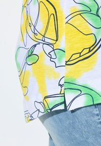 Camicetta a maniche corte con una base bianca, motivo astratto giallo e verde, con contorni neri e dettagli con spacco laterale.