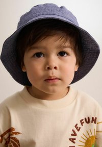 Blauer Bucket Hat mit strukturiertem Finish, kombiniert mit einem beigen Pullover, der ein braunes Blätterdesign und den Text "HAVE FUN" am Kragen trägt.