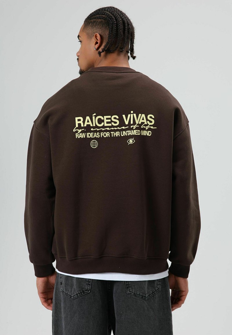 Bruine oversized trui met gele tekst op de achterkant: "RAÍCES VIVAS" en "RAW IDEAS FOR THE UNTAMED MIND." Zachte textuur en geribbelde manchetten.