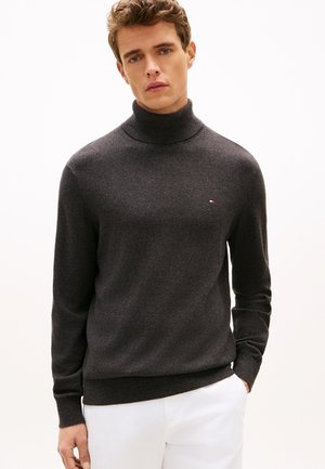 Jeune homme portant un pull à col roulé gris foncé et un pantalon blanc, debout devant un fond clair uni.