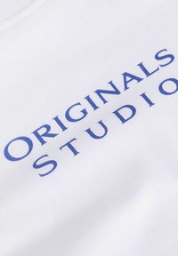 Lettres majuscules bleues "ORIGINALS STUDIO" imprimées sur un tissu blanc, montrées sous un angle diagonal rapproché.