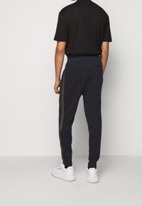 Black sweatpants with a gold logo print along the side, made of soft fabric. Paired with white sneakers, featuring a relaxed fit and elastic waistband.