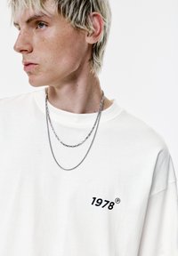 Hvid bomulds t-shirt med en sort "1978" print på venstre bryst. To sølv kæder hænger om halsen. Struktureret stof og afslappet pasform.