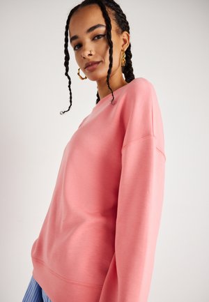 Rosa Sweatshirt mit langen Ärmeln, rundem Halsauschnitt und lockerem Schnitt. Weicher Stoff, minimale Nähdetails, getragen über blauen gestreiften Hosen.