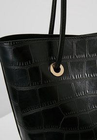 Sac fourre-tout noir avec un motif crocodile en relief brillant. Il présente de longues poignées noires et un accent en métal doré rond.