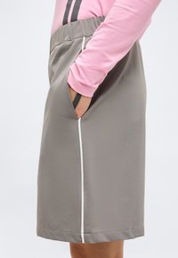 Άτομο που φοράει γκρι φούστα με λευκή πλαϊνή ρίγα και ελαστική μέση, με το χέρι στην τσέπη, συνδυασμένη με ροζ μπλούζα μακρυμάνικη.