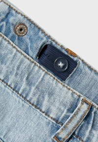 Jean en denim bleu clair avec un bouton en laiton à la taille et des détails cousus, y compris une pièce en tissu bleu marine et des coutures texturées.