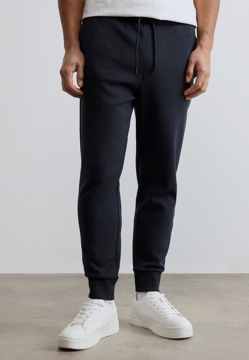 Άνδρας που φοράει μαύρο παντελόνι jogging με ριγμένα μπατζάκια, λευκά sneakers χαμηλού τύπου και λευκό πουκάμισο, στέκεται σε λείο πάτωμα.