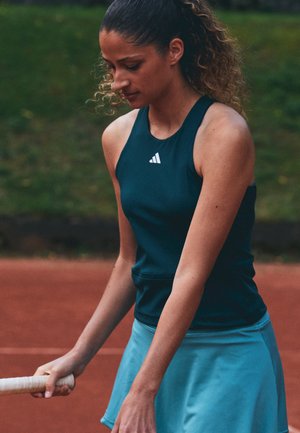 Joueuse de tennis portant un vêtement de sport bleu, tenant une raquette, debout sur un court de terre battue avec un arrière-plan vert.
