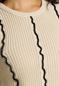 Krems krāsas adīts džemperis ar ribotu apaļu kaklu, vertikālām melnām viļņainām svītrām un ar teksturētu audumu papildu dimensijas piešķiršanai.