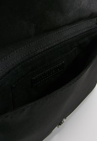 Intérieur d'un sac  à main en tissu noir présentant une texture lisse, des coutures cousues et une petite poche avec une étiquette de marque. Aucun élément métallique visible.
