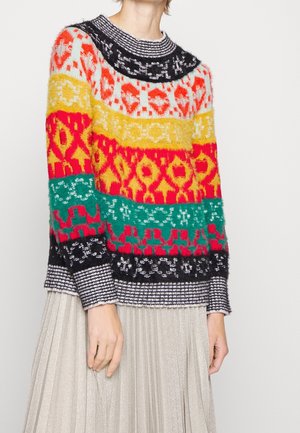 Personne portant un pull en tricot à motifs multicolores avec des rayures rouge, jaune, vert, noir et blanc et une jupe plissée argentée-grise.