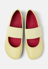Gula platta skor med ett gummiliknande övre, rund tå och bred elastisk rem. Inuti finns en kontrasterande röd innersula.