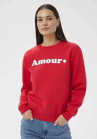 Femme aux longs cheveux bruns portant un sweat-shirt rouge avec "Amour" et un cœur imprimés en blanc, mains dans les poches d'un jean bleu, fond neutre.