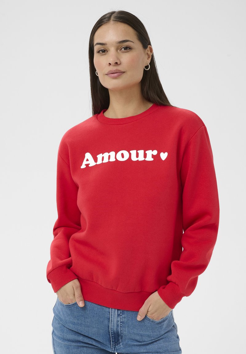 Femme aux longs cheveux bruns portant un sweat-shirt rouge avec "Amour" et un cœur imprimés en blanc, mains dans les poches d'un jean bleu, fond neutre.