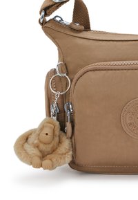 Borsa a tracolla in tessuto marrone con scomparti con cerniera. Presenta un logo circolare e una spilla a forma di scimmia in peluche attaccata come dettaglio.
