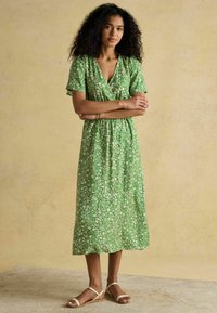 Grünes Blumenkleid mit kurzen Ärmeln, V-Ausschnitt und tailliertem Bund. Hergestellt aus leichtem Stoff mit einem mittellangen Saum. Dazu werden flache Sandalen getragen.