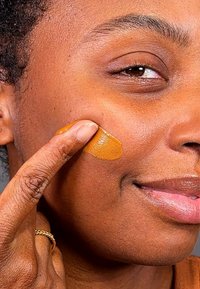 Brun hud med en slät yta, där en finger applicerar en gyllenbrun ansiktsprodukt på kinden. Naturligt ljus framhäver huden.